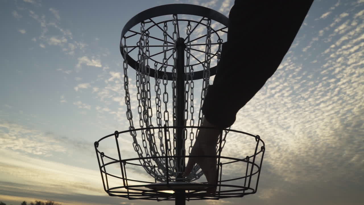 Man plockar upp en discgolf-frisbee från en discgolfkorg silhuetterad mot skymningshimlen