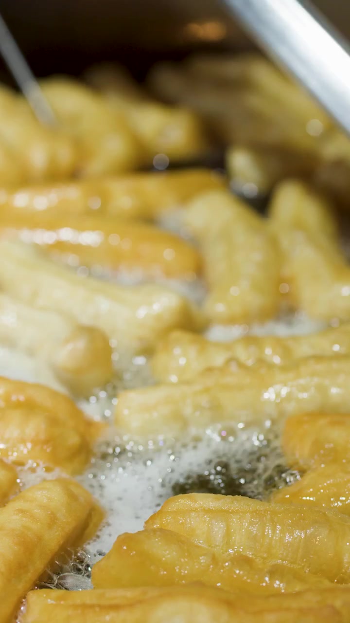 Primer plano de youtiao dorados friéndose en aceite burbujeante con pinzas metálicas dándoles la vuelta.