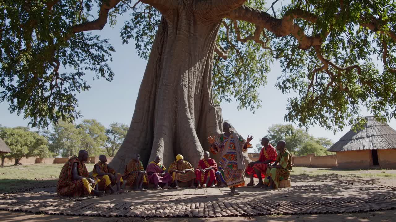 Wide-angle video captures a group seated under a large tree, dressed in colorful attire