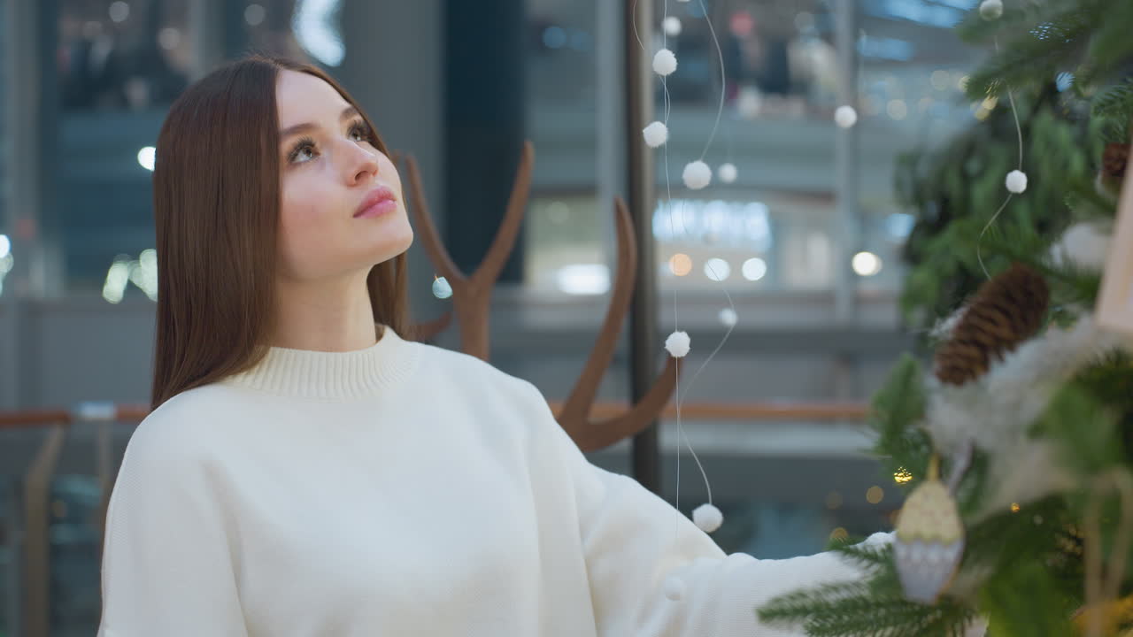 Elegant woman in white sweater gazes warmly at festive decor in beautifully decorated mall, surrounded by Christmas ornaments, glowing fairy lights, and a serene holiday atmosphere