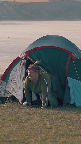 pudgy girl with hard hangover crawls from blue tent holding metal cup on pictorial river steep bank slow motion