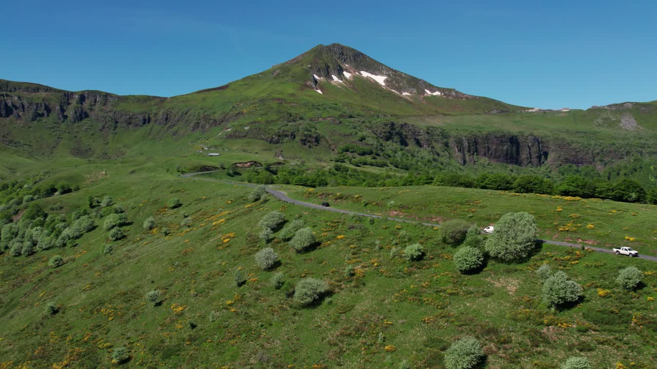 Aerial shot of a campervan driving on a roas in Cantal with the Puy Mary Volcano in the background on a beautiful summer day, Cantal departement, Auvergne region, France