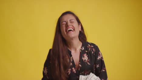 studio shot of excited woman celebrating winning cash prize throwing handful of 100 dollar bills in the air against yellow background