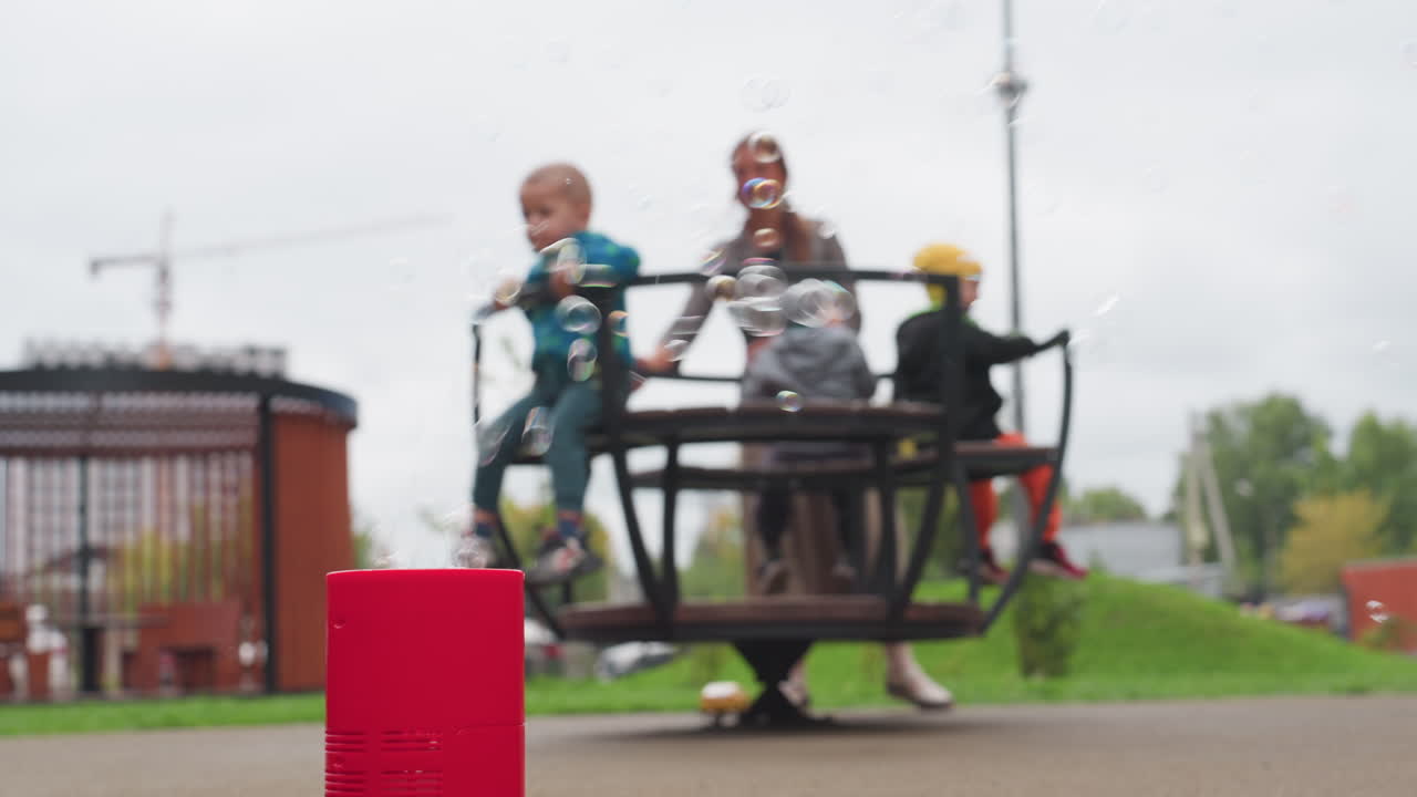 Una donna fa girare dei bambini su una giostra mentre delle bolle salgono da una macchina rossa in primo piano, parco giochi con messa a fuoco morbida, prato verde, cielo nuvoloso, giro leggero e atmosfera gioiosa in un parco urbano