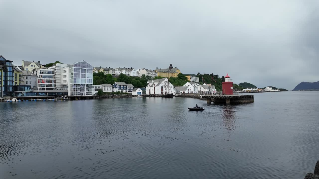 Norveç, Ålesund'un geleneksel beyaz binaları, kırmızı bir deniz feneri, sakin liman suları ve sisli dağlarla dramatik kapalı gökyüzünü içeren manzaralı sahil şeridi görünümü.