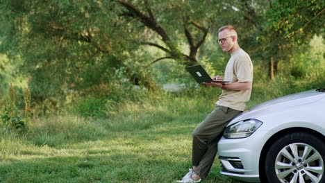 mid-aged man stands leaning on car outdoors. man works on laptop holding it in hands. freelancer takes off glasses looking ahead pensively.