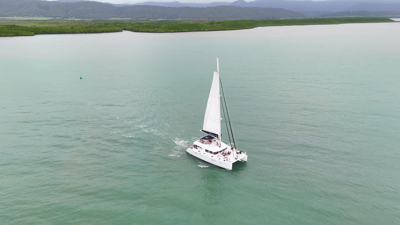 Drone captures a catamaran sailing smoothly in calm, turquoise waters under overcast skies