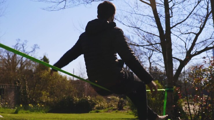Low angle of barefoot young man in jeans and puffer jacket sitting on slack line and balancing