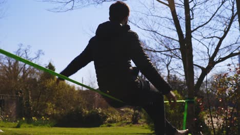 low angle of barefoot young man in jeans and puffer jacket sitting on slack line and balancing