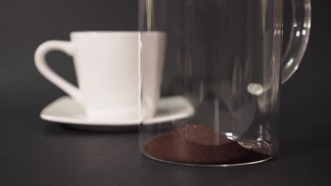 filling french press with ground coffee with a white cup in the background - closeup