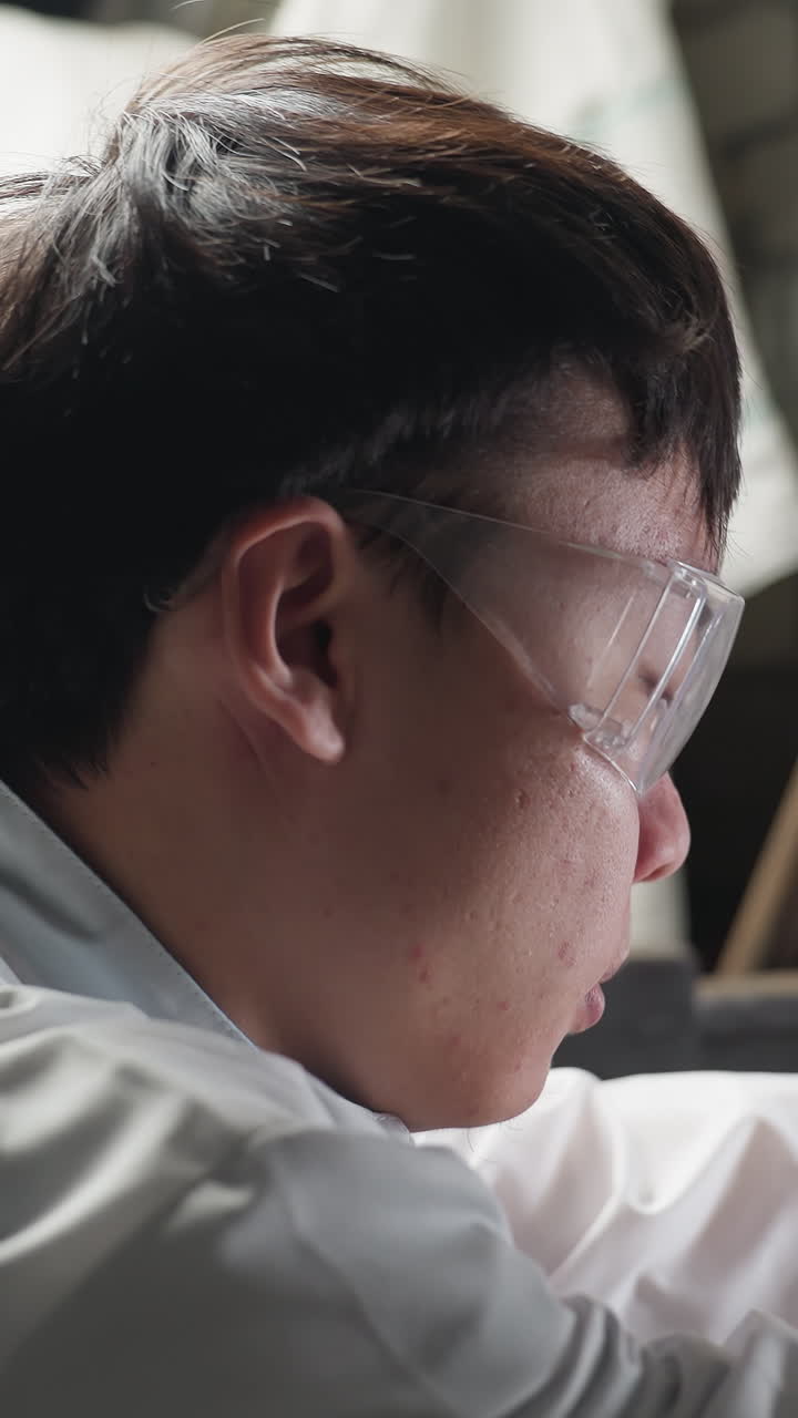 색 실험실 코트를 입은 중국 학생이 자동차 수리 작업장에서 보호 안경과 태블릿을 들고 작업을 준비하며 엔지니어링 설정에서 안전과 현대 기술을 강조합니다.