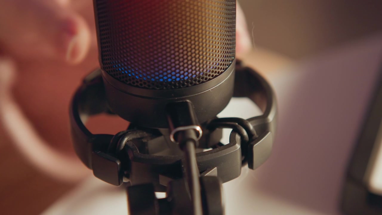 Close-up shows hands tightening shock mount around large studio microphone while blue and magenta LED glow reflects across mesh surface, capturing careful adjustment, secure grip, and recording