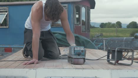 Young-carpenter-wearing-white-vest-belt-sanding-cabin-roof-planking-of-old-wooden-boat