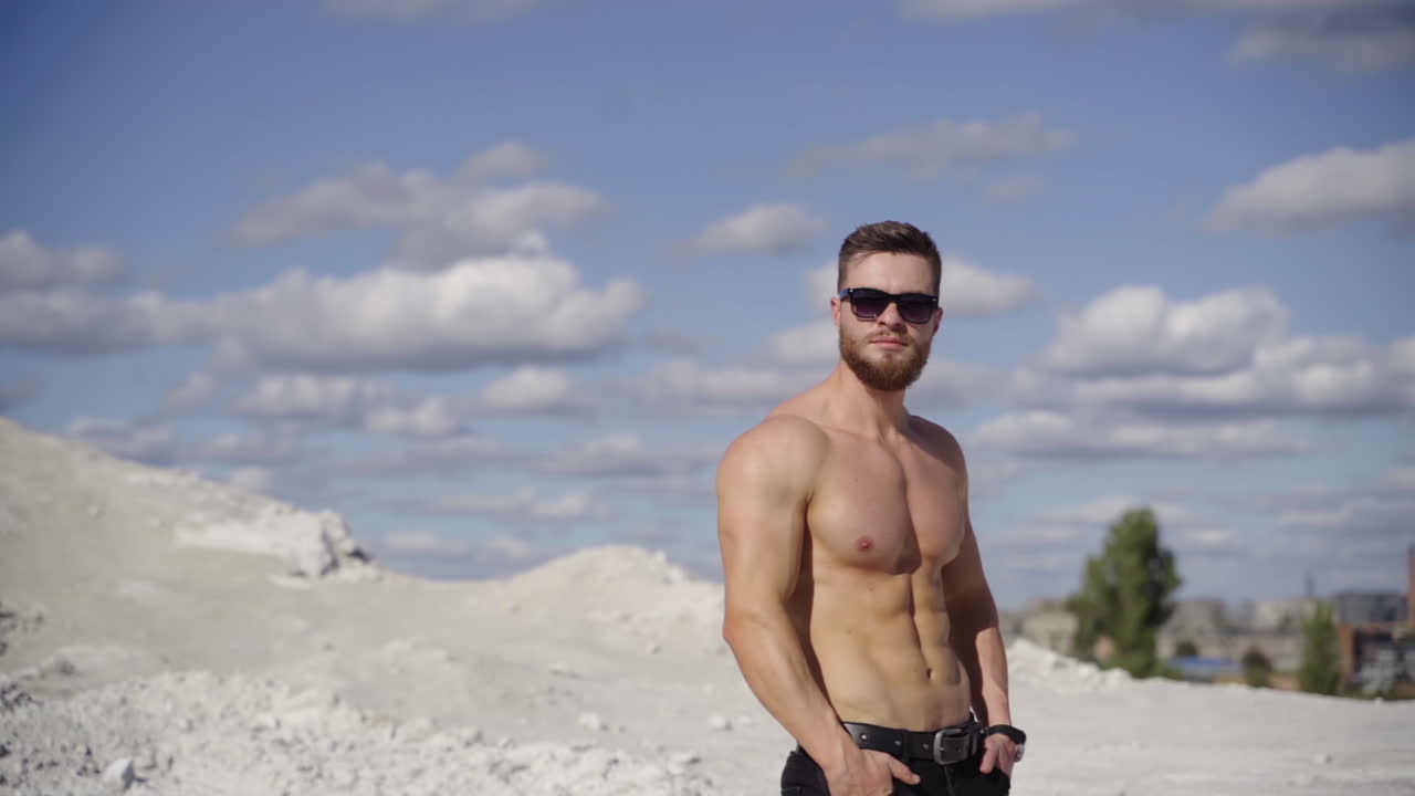 Portrait of athlete at sunlight. Handsome shirtless man in black sunglasses standing on the top of white hill against the beautiful summer sky. Healthy lifestyle.