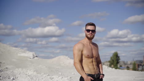 portrait of athlete at sunlight. handsome shirtless man in black sunglasses standing on the top of white hill against the beautiful summer sky. healthy lifestyle.
