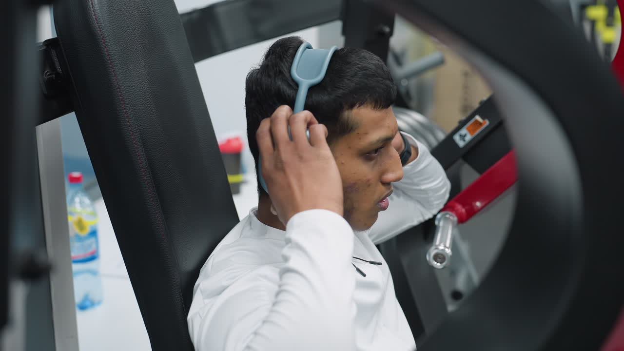 close up of gym coach sitting on workout machine while adjusting wireless headphones before starting exercise session inside fitness center, focused and determined with background