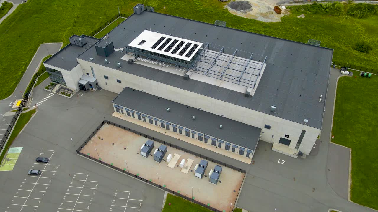 Aerial drone footage ascending higher before and infront of a large modern gray colored data center or power station warehouse during a summer cloudy day with green grass around. Solar panels on top