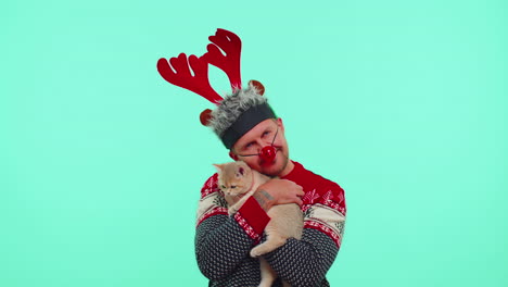 encantador hombre adulto en el suéter de navidad de moda y el sombrero de cuernos de ciervo sosteniendo su querida mascota gato