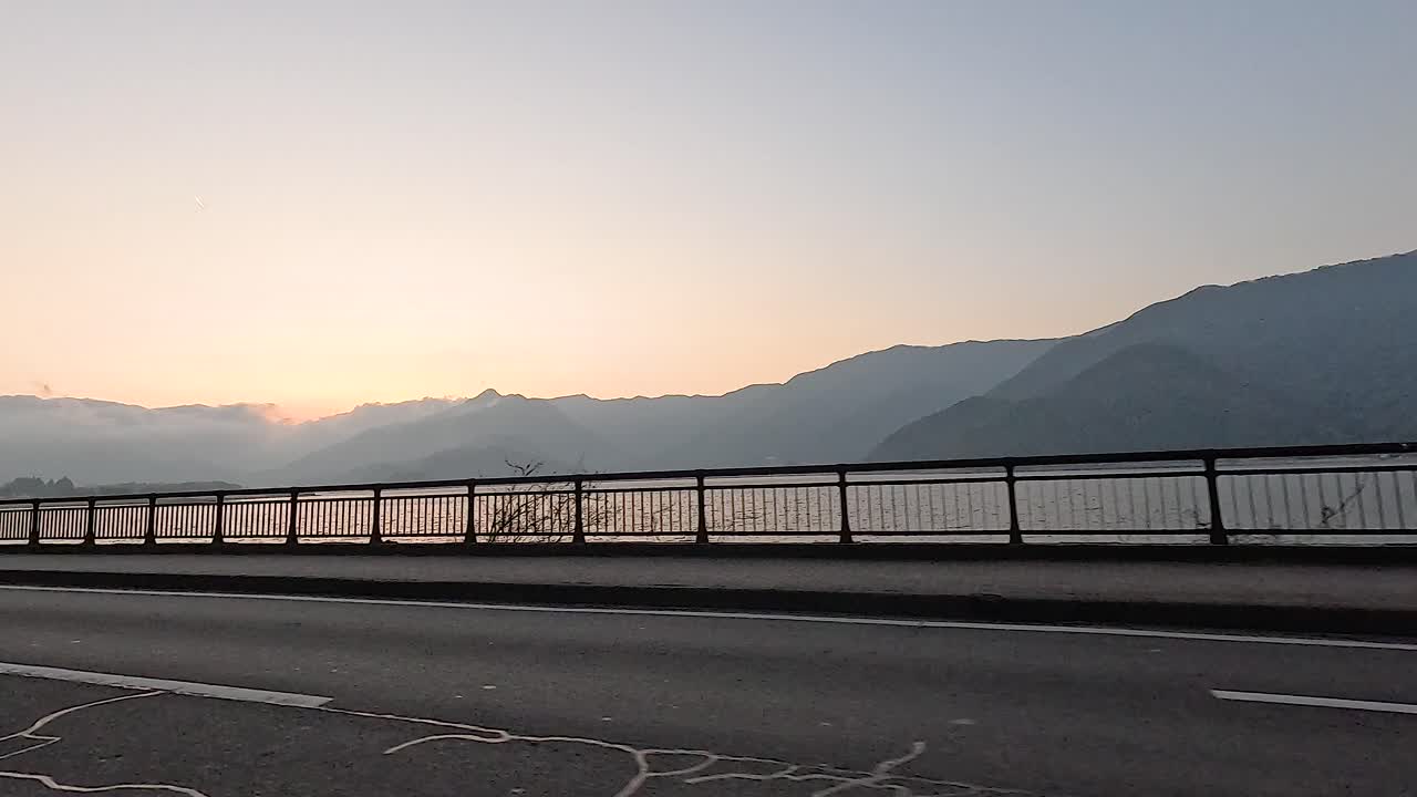 Cars drive along a serene mountain road at sunset near Fujikawaguchiko, Japan. The tranquil scene features silhouetted mountains and a calm lake