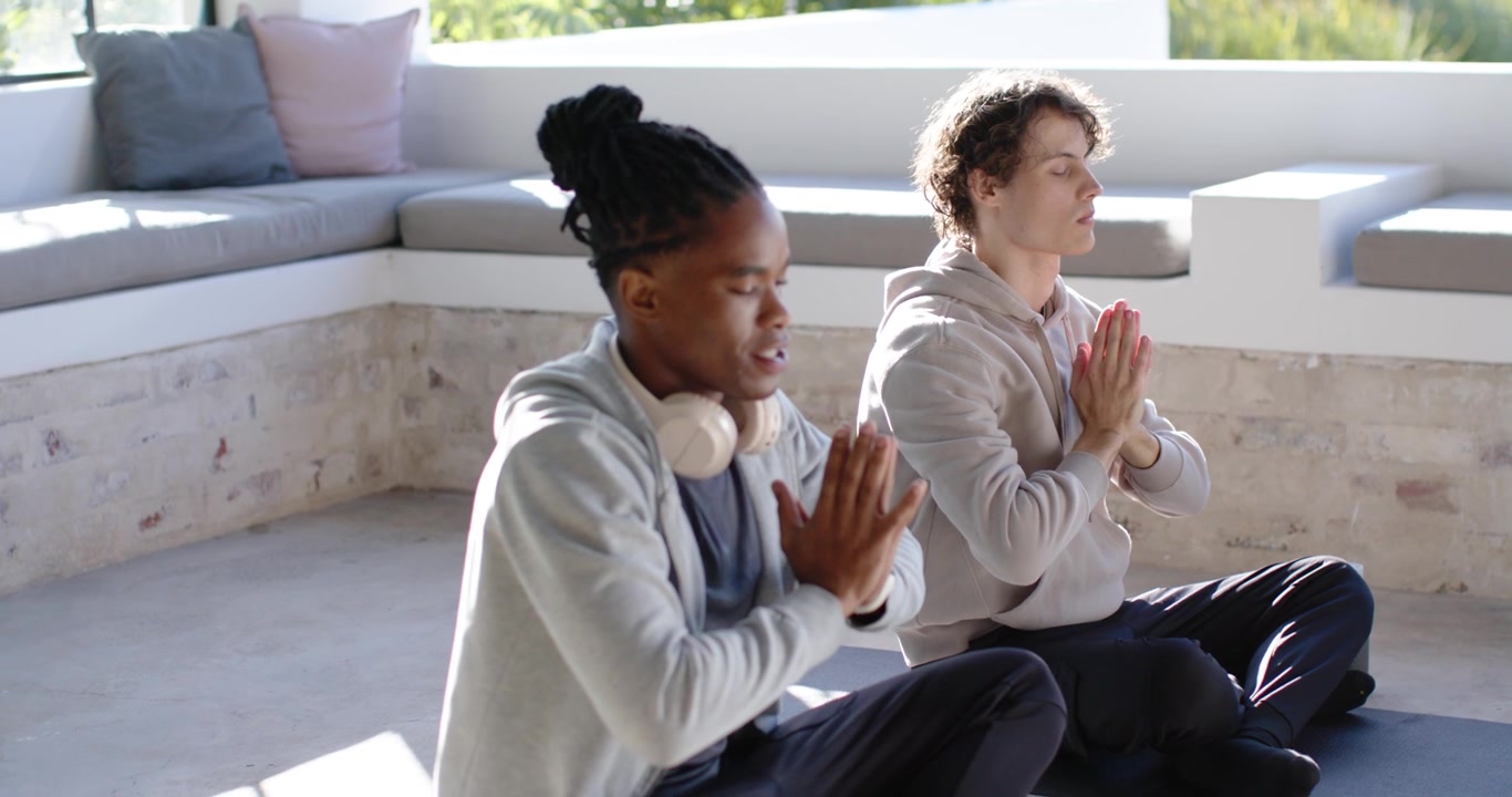 Diverse men following breath cue, raising arms, joining palms, closing eyes on mats in studio