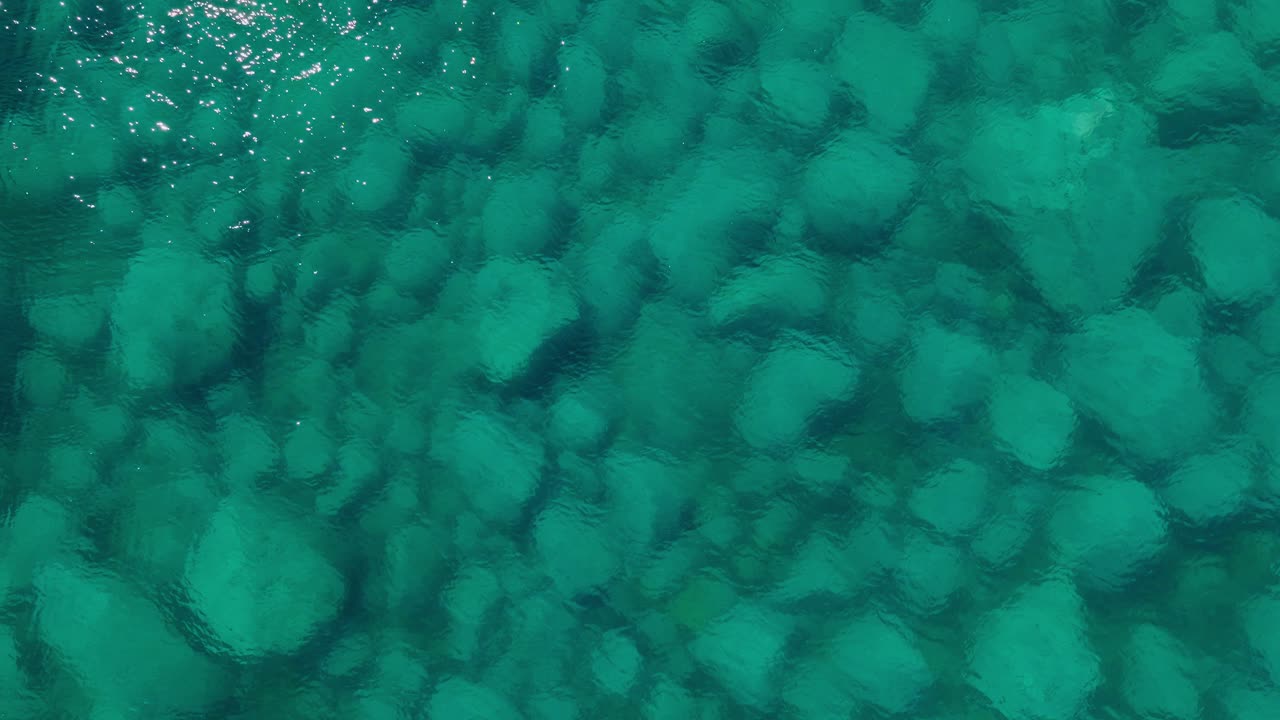 Aerial tilts up above clean sea water revealing stones, natural landscape with sunshine reflection