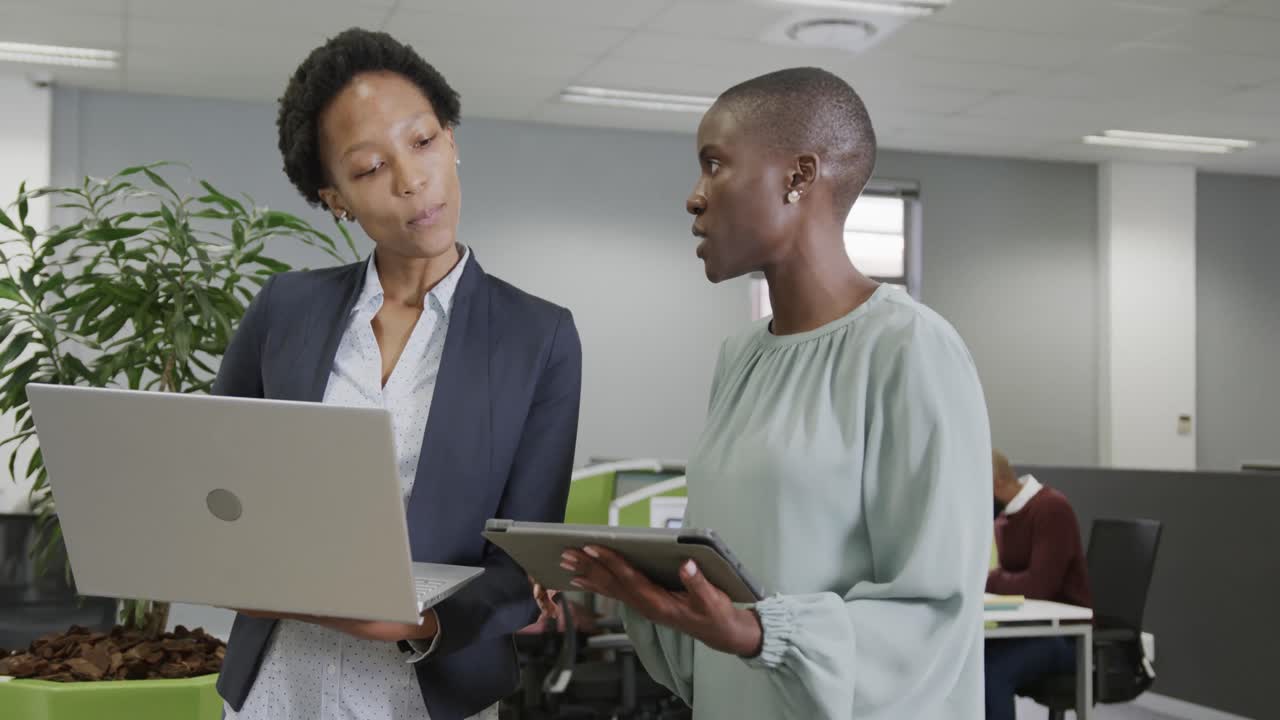 Due donne d'affari afroamericane discutono di lavoro, utilizzando un laptop e un tablet in ufficio al rallentatore.