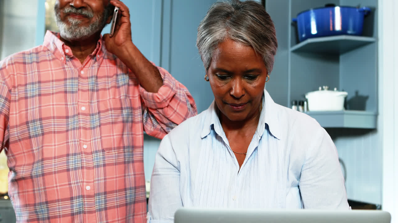 Coppia anziana che usa il cellulare e il laptop in cucina
