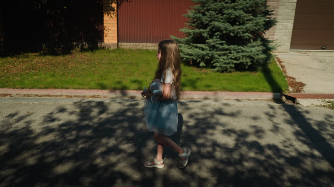 Little girl walks slowly under tree shade on residential street holding her gown with one hand and plush toy in other, sunlight filtering through branches creating soft patterns on asphalt