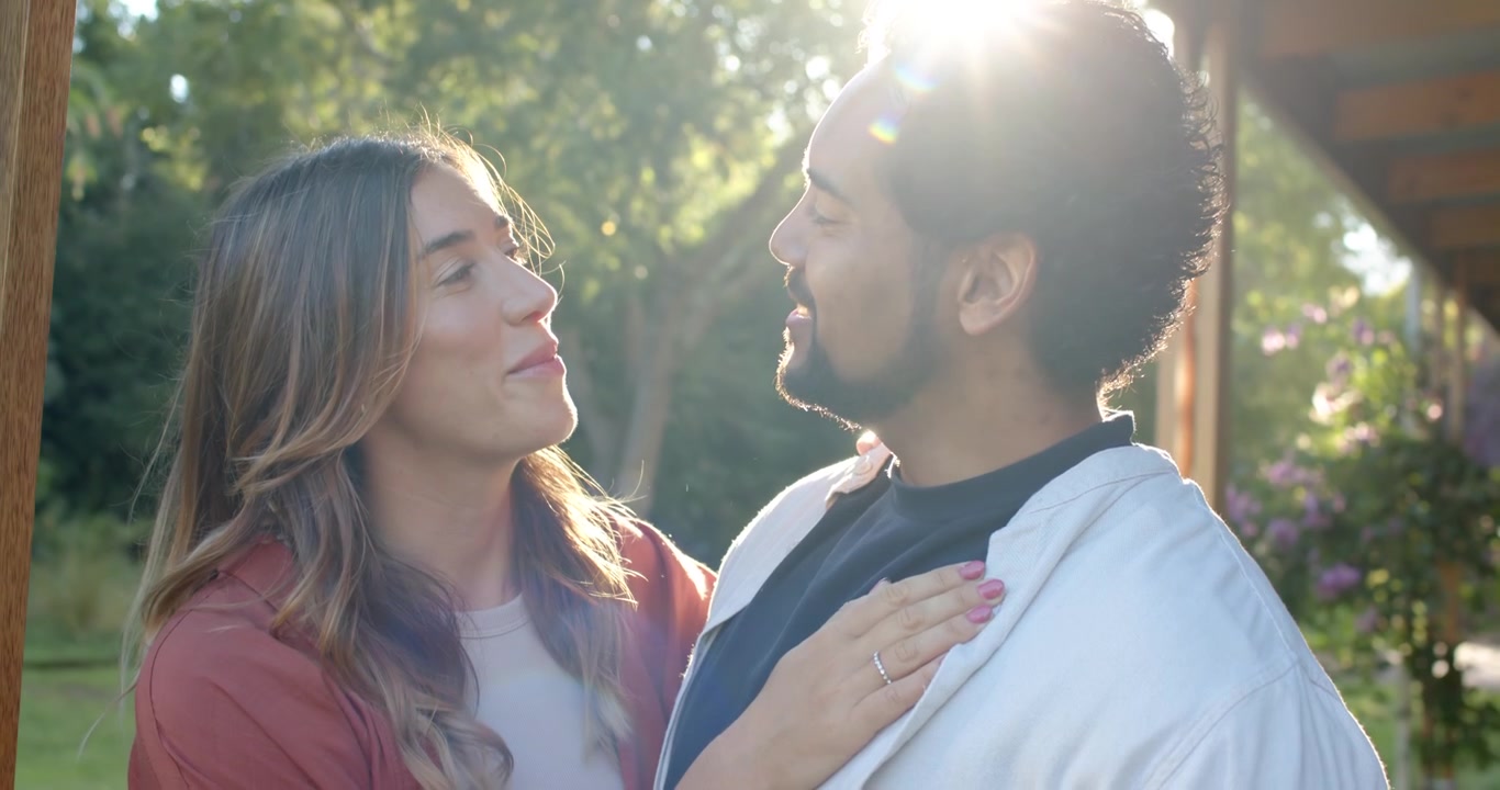 Pareja de pie en el borde del porche junto a un poste de madera, observando un destello solar, mirándose y abrazándose estrechamente.