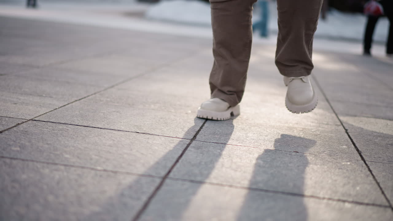 Closeup of woman legs in beige corduroy pants and chunky white platform shoes moving around on snow dusted tiled plaza ground under pale winter sunlight casting long shadows and adding rhythm