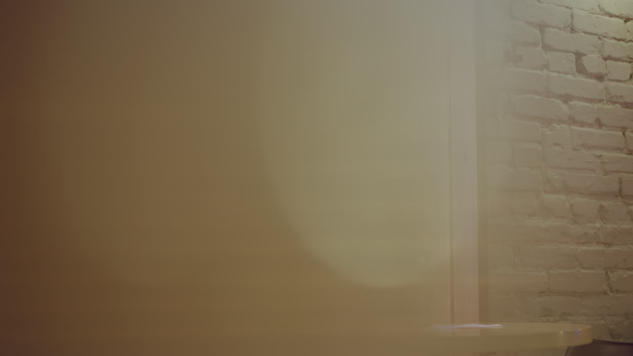 Joven con camisa casual sentado solo en una mesa de cafetería cerca de una gran ventana con vista a la ciudad, sosteniendo un vaso de papel vacío con pajita, rodeado de luces de espejo, capturado desde atrás en un tranquilo entorno interior.
