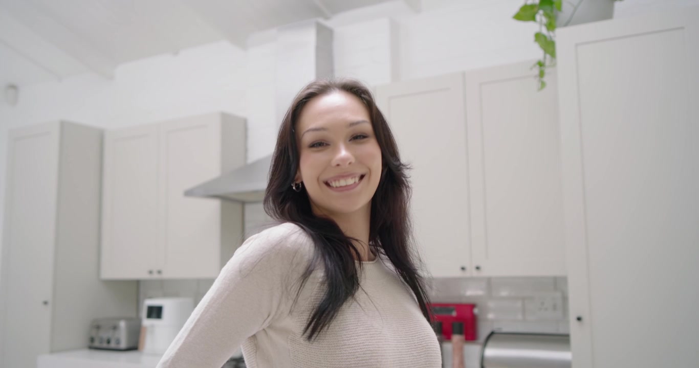 Donna adulta che guarda la telecamera, in posa con le mani sui fianchi e sorridendo in una cucina di casa con cappa aspirante.