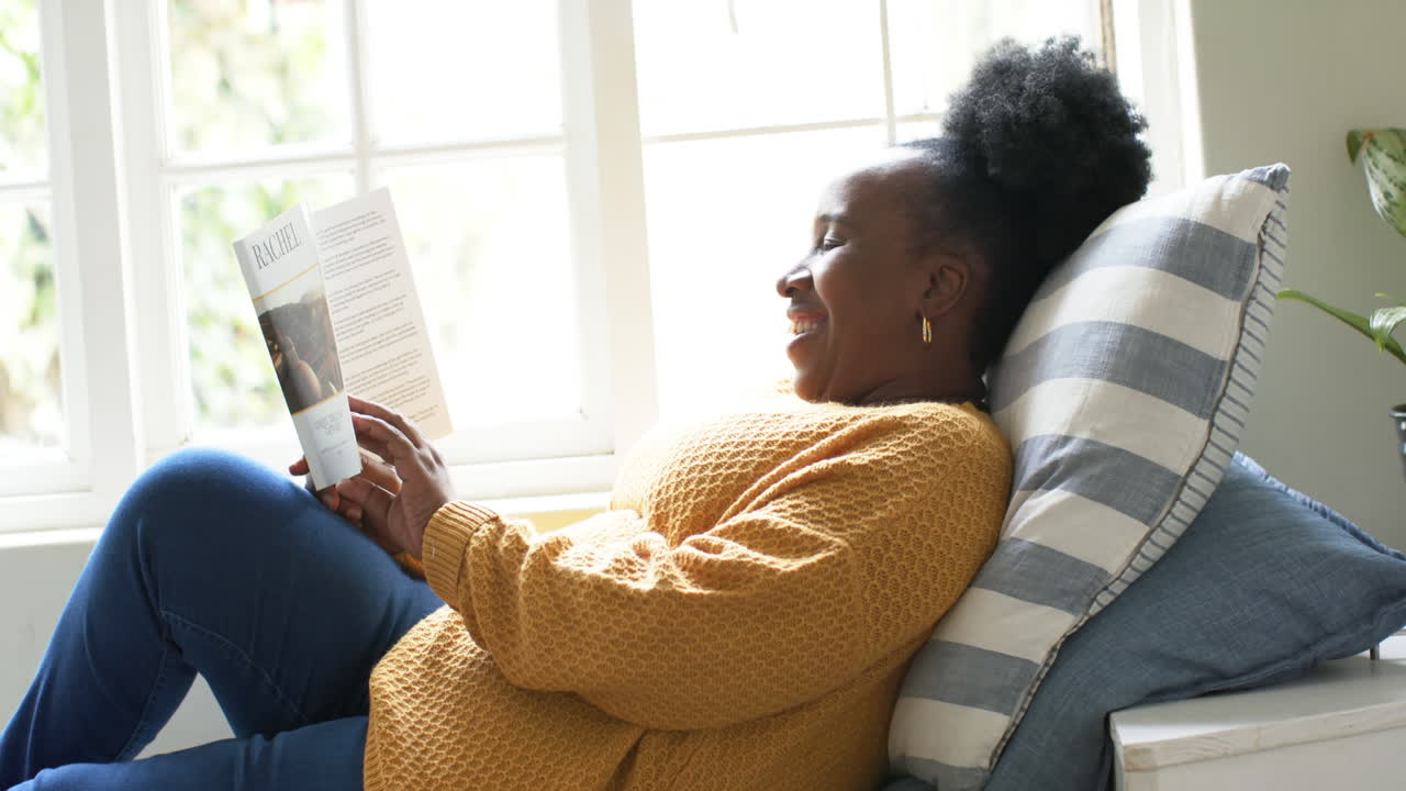 Happy african american senior woman lying, reading book and smiling in sunny room, slow motion