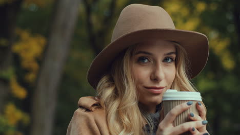 close-up view of caucasian young blonde woman in a hat drinking hot coffee sitting on a bench while looking at the camera in the park in autumn