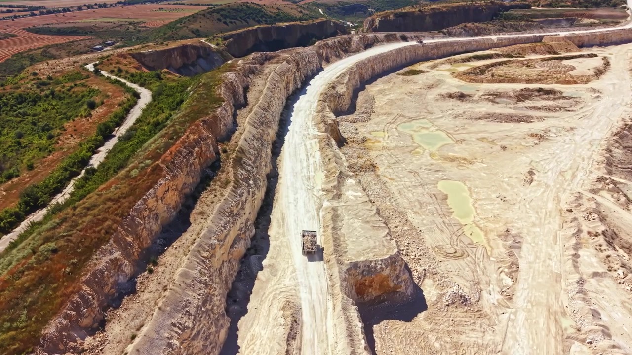 Cava che mostra movimento terra e funzionamento di macchinari in un paesaggio con rocce stratificate e vegetazione distante.