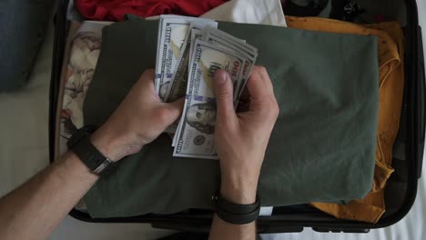 Unrecognizable-man-hands-with-pile-of-money,-US-dollars-notes---counting-under-the-suitcase-with-clothes.-Counting-money-for