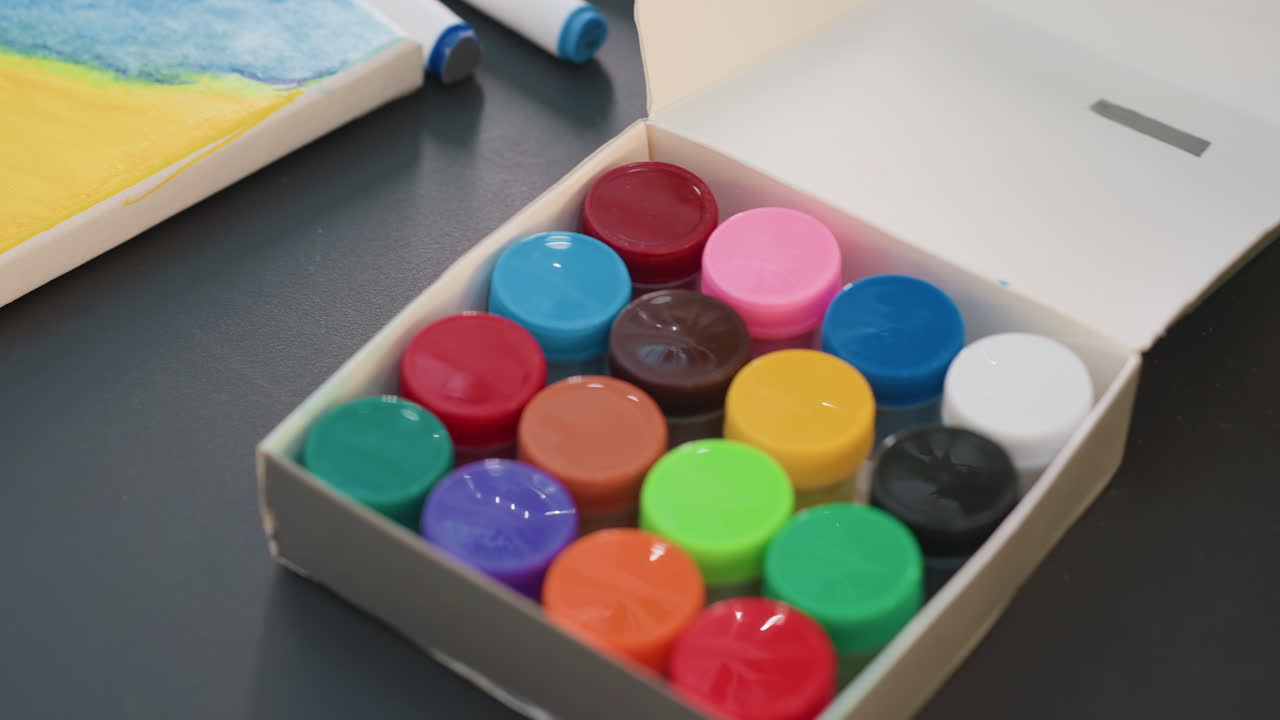 Tarros de pintura de colores en una caja sobre un escritorio, primer plano de pequeños botes de acrílico dispuestos en forma de cuadrícula, rotulador y bloc de acuarelas junto a la caja, la iluminación del estudio resalta las tapas de plástico brillante, rica variedad de pigmentos evoca