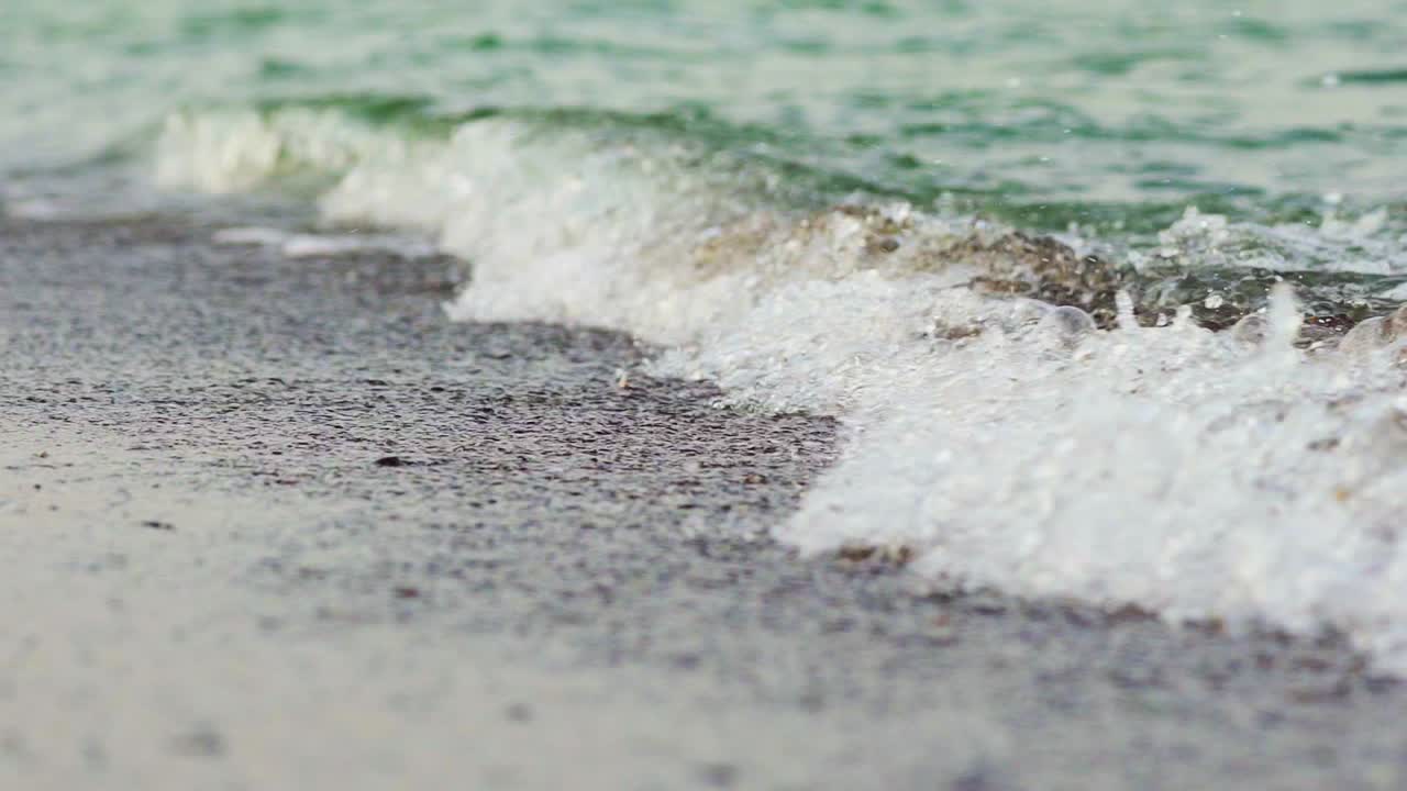 A turquoise sea waves cover the coast and take small grains of sand with it. Close-up. Blurred background.