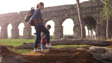 beautiful young woman backpacker walking balancing on a log trunk holding hands with man boyfriend in front of ancient roman aqueduct ruins in parco degli acquedotti park in rome at sunrise romantic sleeping bag slow motion