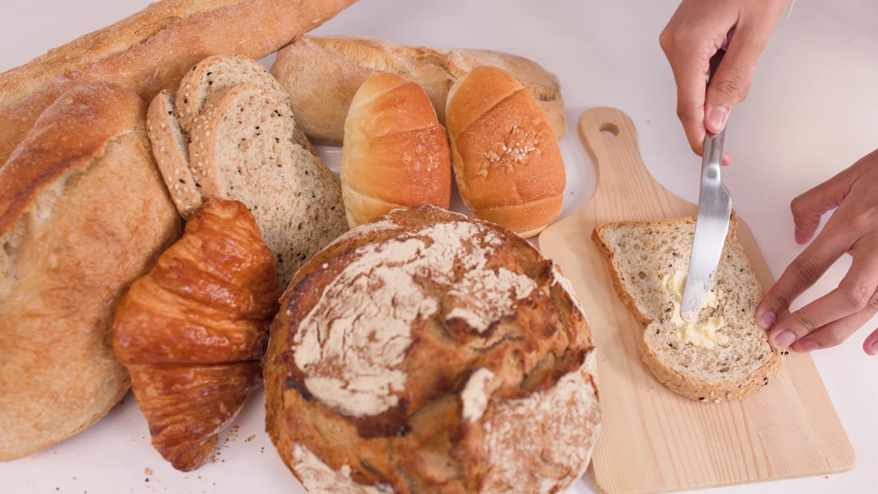 Primo piano di mani che spalmano burro su una fetta di pane, circondate da pane a lievitazione naturale, baguette e croissant.