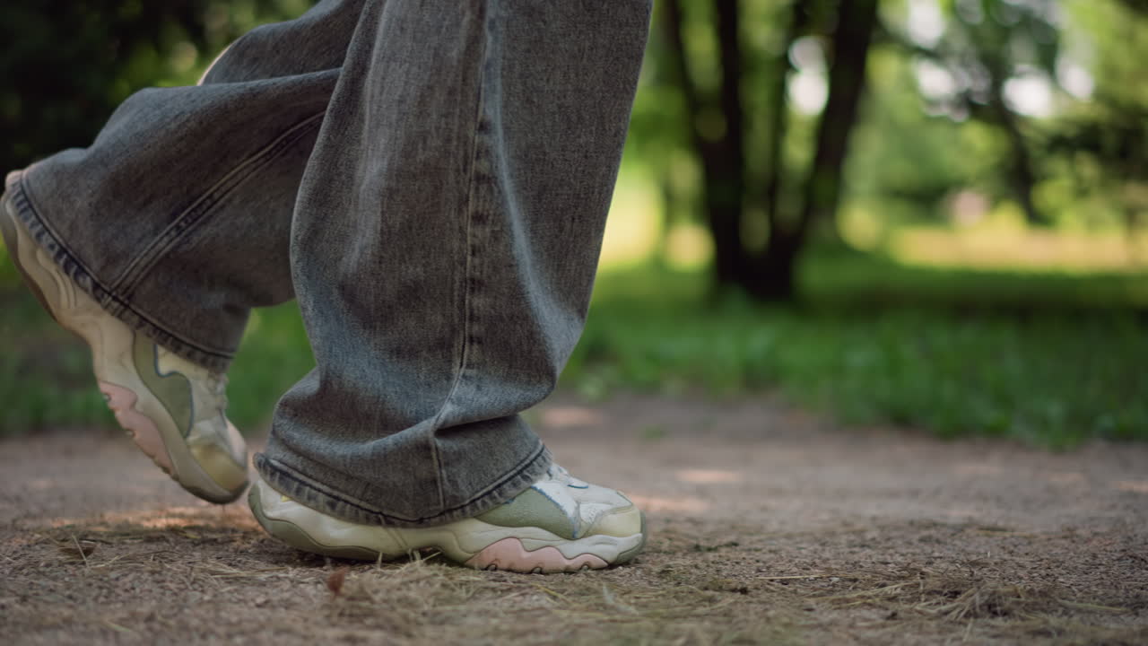 Sentiero del parco, primi piani di piedi che camminano lungo la ghiaia sotto gli alberi, jeans casual e scarpe da ginnastica usurate catturano la luce solare a chiazze, passo lento e ritmico che solleva una leggera polvere, trasmettendo calma, radicamento, ritmo meditativo