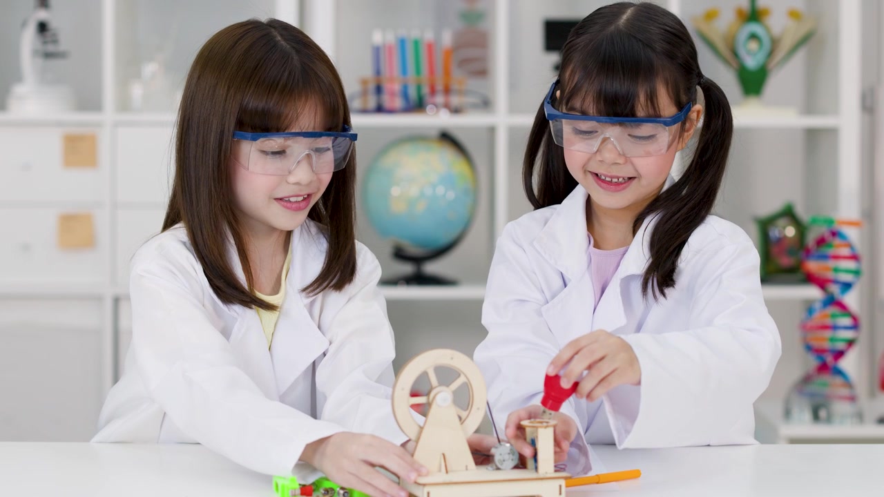 Two schoolgirls wearing safety goggles and lab coats assemble a wooden STEM project in classroom.