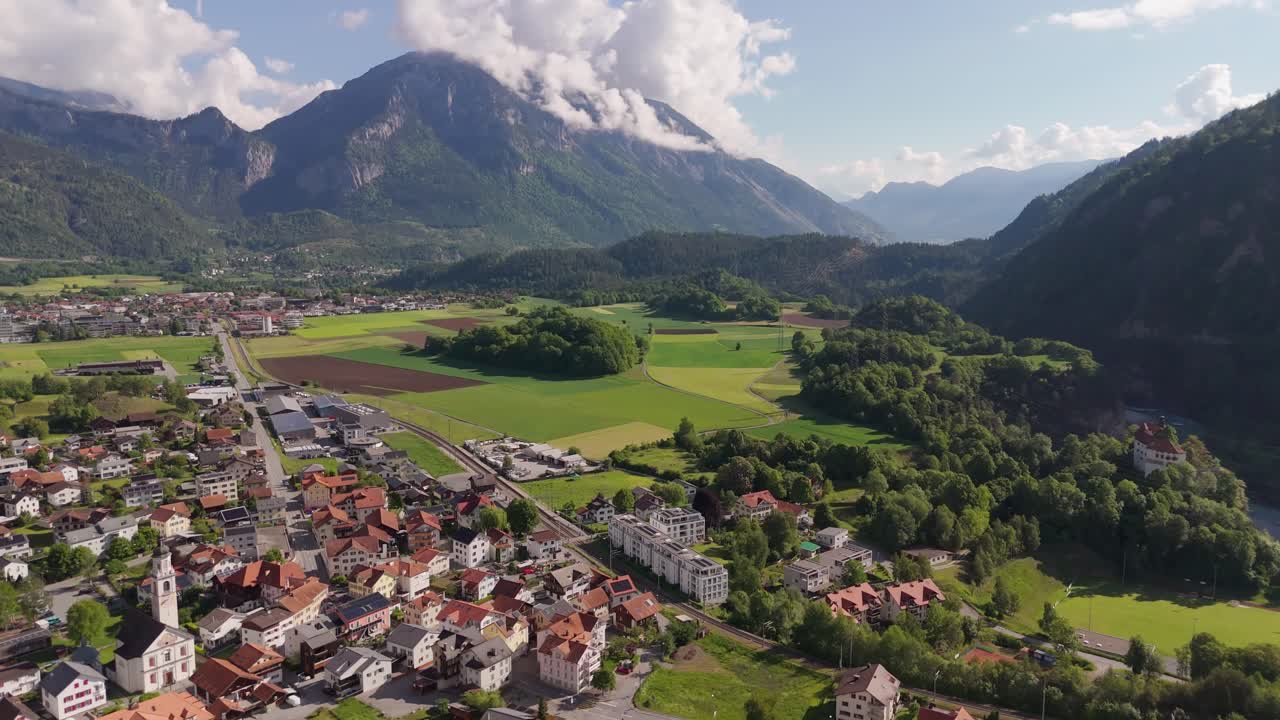 Aerial wide shot of historic houses and Apartments with green farm fields. Rhäzüns Swiss City during sunny day and alps in distance. Beautiful spring day with green trees in Switzerland.