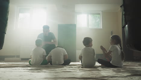 trainer talks to kids at break in martial arts club. little children rests with young instructor sitting on floor carpet in gym slow motion. fighting lesson