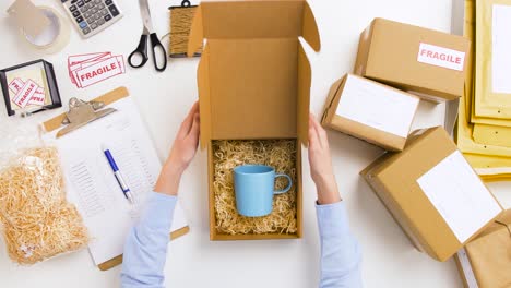 Woman-Packing-Mug-To-Parcel-Box-at-Post-Office-9.delivery,-mail-service,-people-and-shipment-concept-woman-wrapping-mug-into-protective-bubble-wrap-and-packing-it-to-parcel-box,-then-crossing-item-off-form-on-clipboard-at-post-office