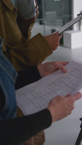 Vertical-Video-Close-Up-Of-Female-Film-Director-And-Assistant-Looking-At-Script-Shooting-Movie-In-Studio-Shot-In-Real-Time