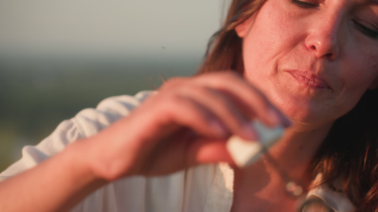 Young woman blowing soapy bubble wand with eyes closed and slight smile, bathed in warm sunset light on grassy hillside overlooking tranquil river valley, gentle breeze moving hair