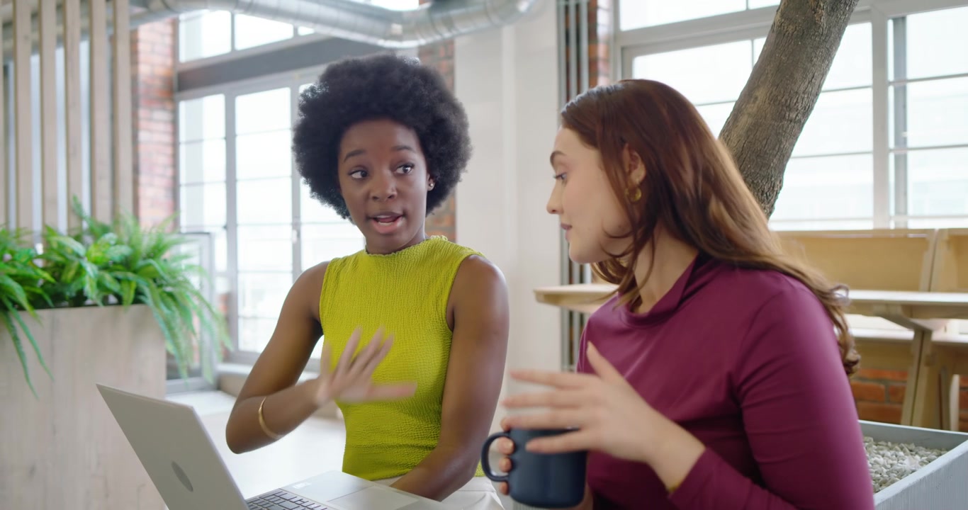 Diverse female coworkers leaning and discussing work on laptop in office with mug after focus pull