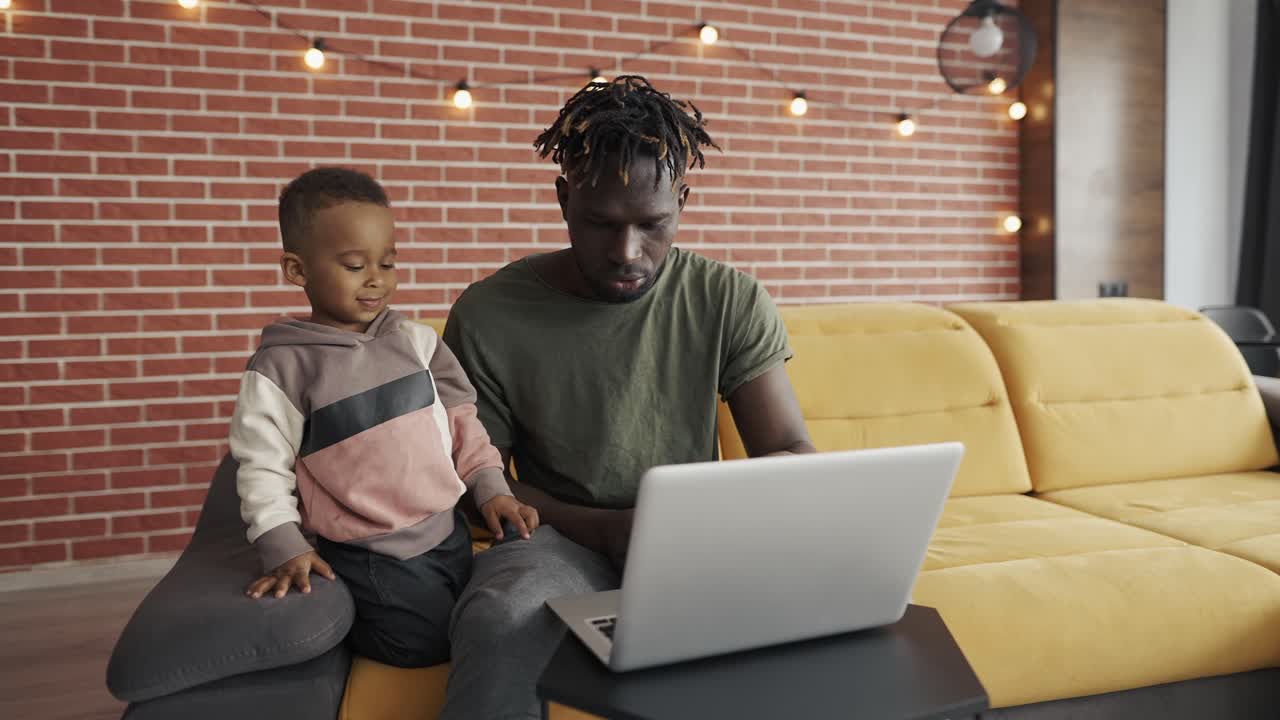 Father and small boy typing together on laptop at home on sofa, slow motion