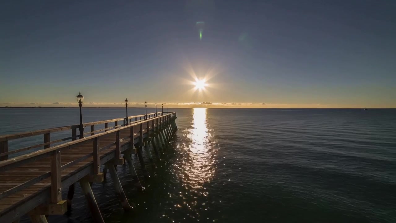 Muelle de madera que se extiende hacia el mar con barandillas y farolas, el sol saliendo e iluminando el agua